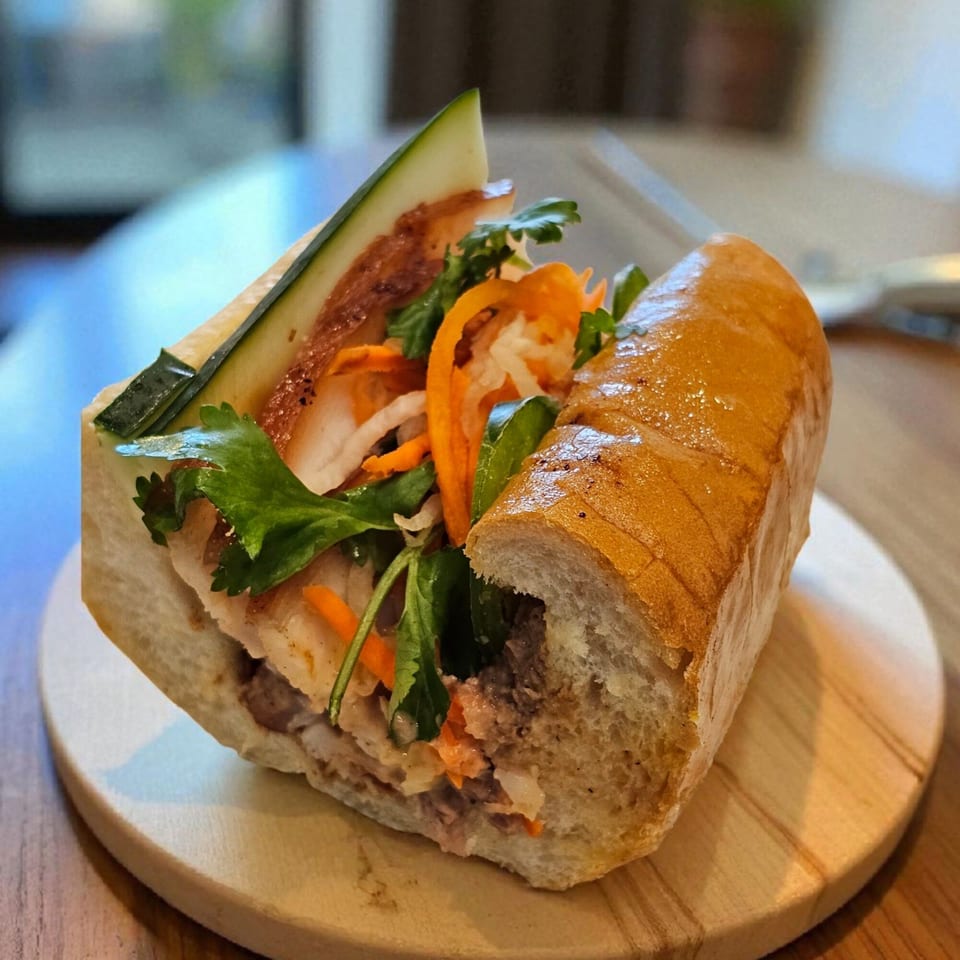 A Vietnamese banh mi sandwich with meat, fresh vegetables, and herbs on a wooden board.