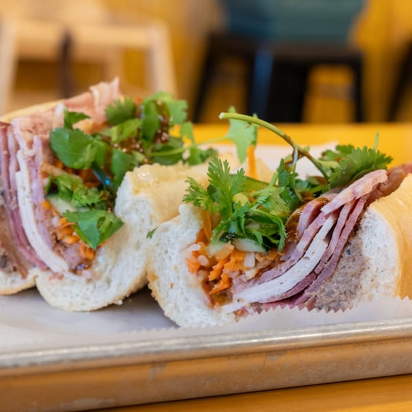 Two halves of a SATE Banh Mi sandwich, filled with meats, pickled vegetables, and cilantro.