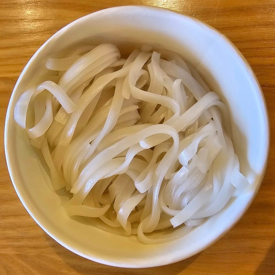 A white bowl filled with flat, white rice noodles, likely for a Vietnamese dish, on a wooden table.