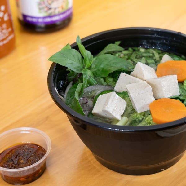 A black bowl of Vietnamese pho with tofu, carrots, broccoli, and basil, with a side of sauce.