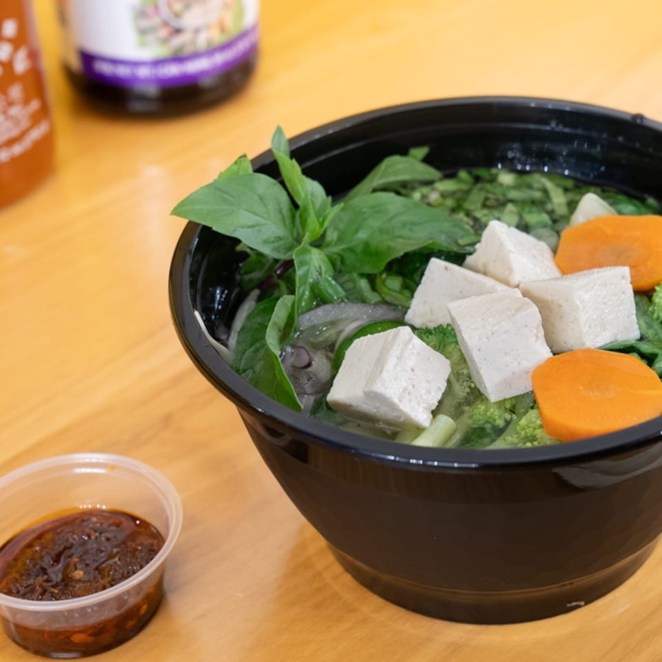 A black bowl of Vietnamese pho with tofu, carrots, broccoli, and basil, with a side of sauce.