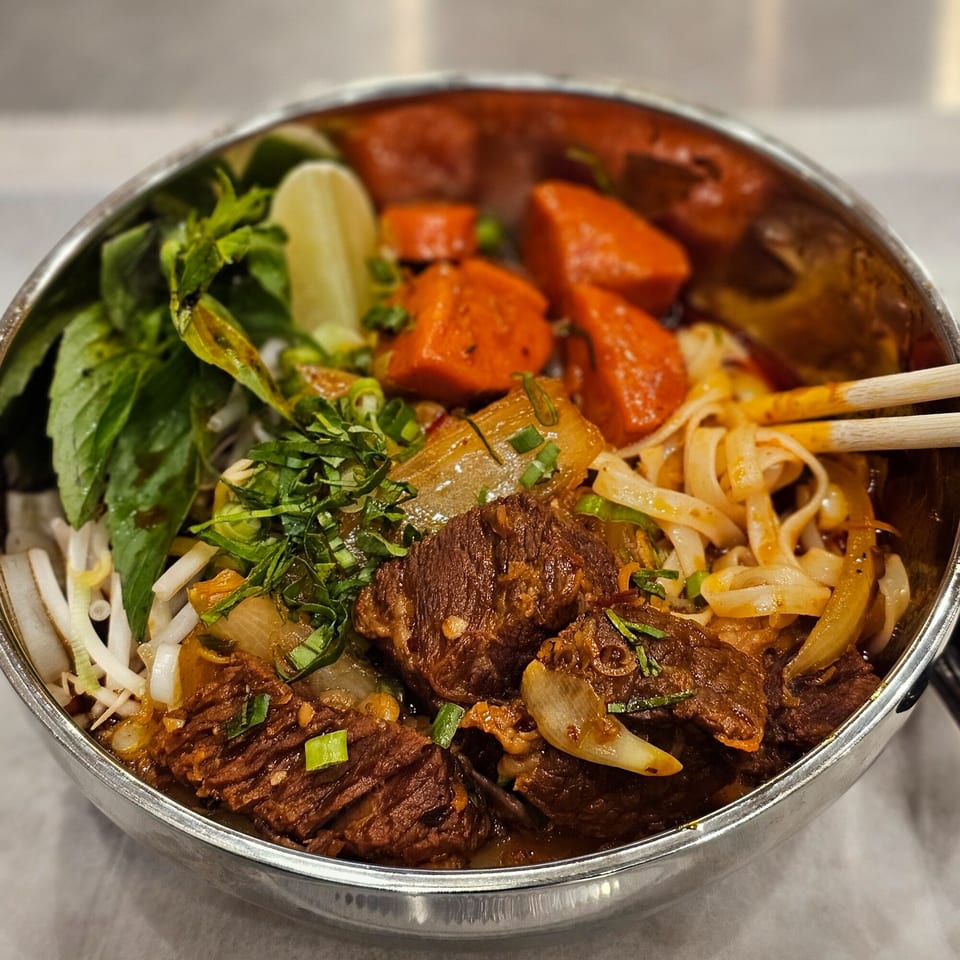 Close-up of a Vietnamese beef stew with noodles, carrots, herbs, and a lime wedge in a metal bowl.