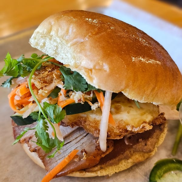 A close-up of a SATE Banh Mi & Grille Vietnamese sandwich with pork, egg, and vegetables.