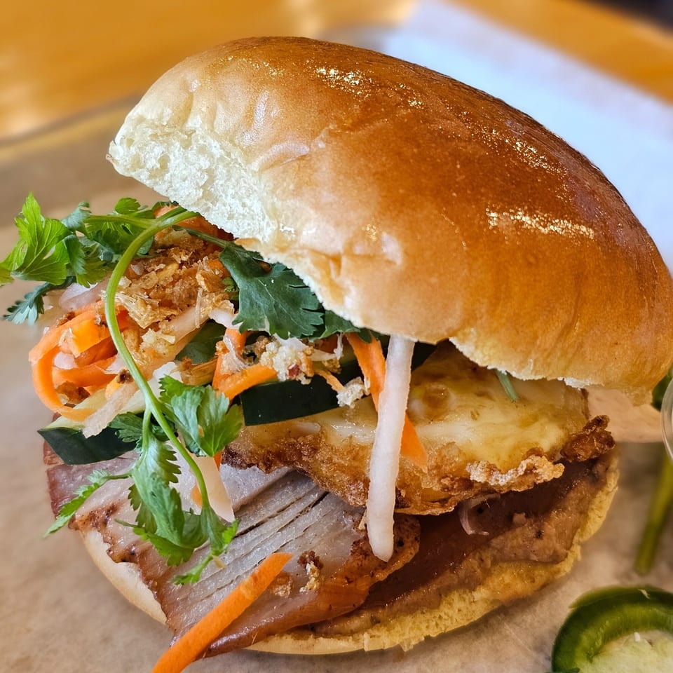 A close-up of a SATE Banh Mi & Grille Vietnamese sandwich with pork, egg, and vegetables.