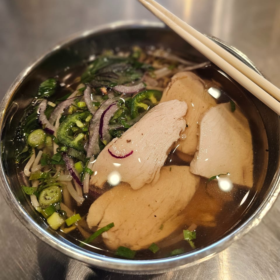 A bowl of Vietnamese pho with chicken, onions, and green herbs, with chopsticks resting on top.