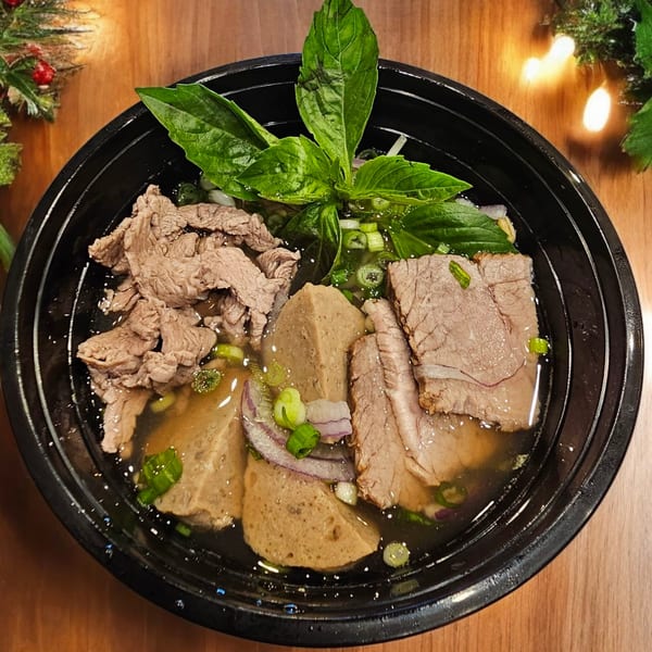 Vietnamese pho with various cuts of beef, meatballs, green onions, and basil in a black bowl.