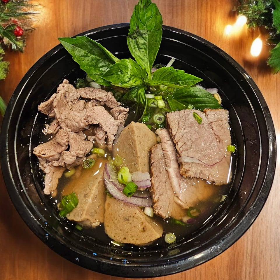 Vietnamese pho with various cuts of beef, meatballs, green onions, and basil in a black bowl.