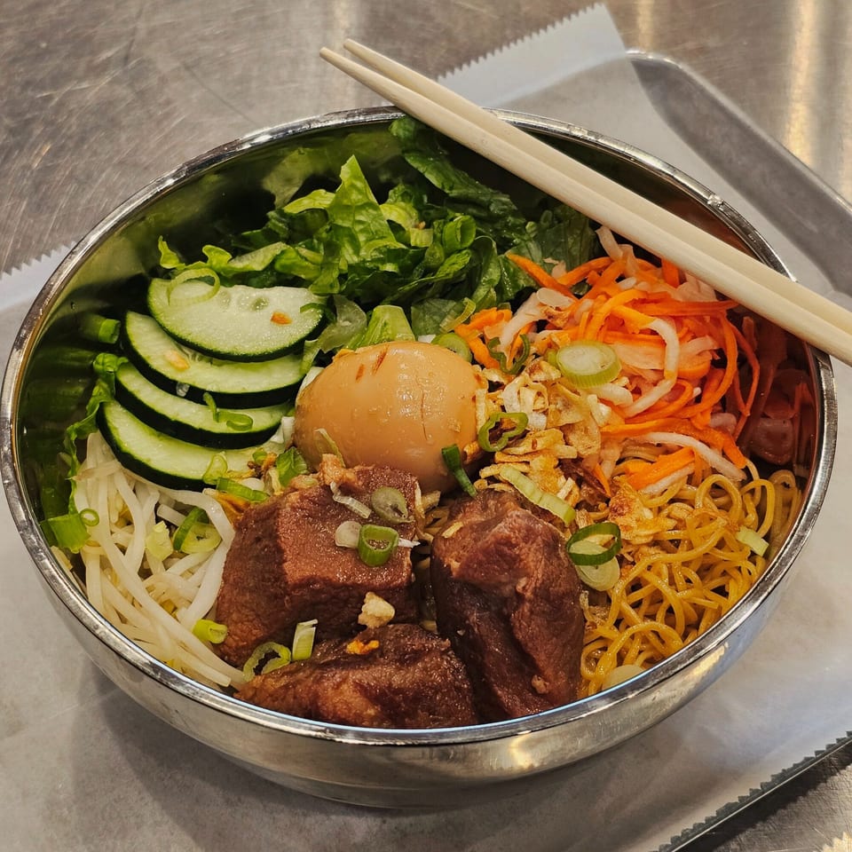 A Vietnamese noodle bowl with beef, egg, vegetables, and chopsticks in a metal bowl.