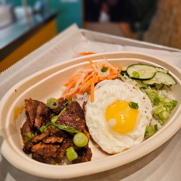 A Vietnamese rice bowl with grilled meat, a fried egg, pickled carrots, cucumbers, and scallions.