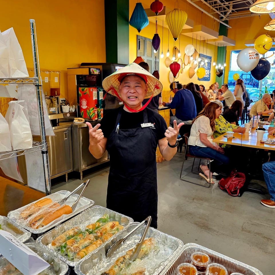 A man in a conical hat and apron stands proudly before Vietnamese food trays and a busy dining area.