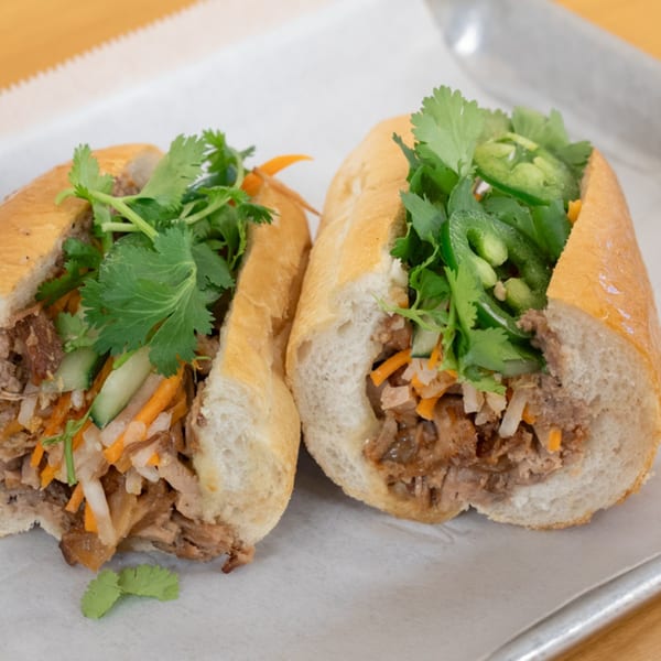 Two SATE Banh Mi sandwiches, cut in half, filled with meat, pickled vegetables, and fresh cilantro.