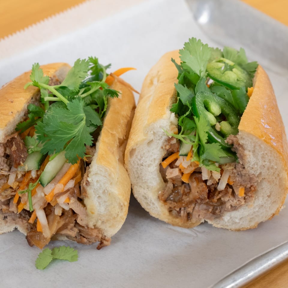 Two SATE Banh Mi sandwiches, cut in half, filled with meat, pickled vegetables, and fresh cilantro.