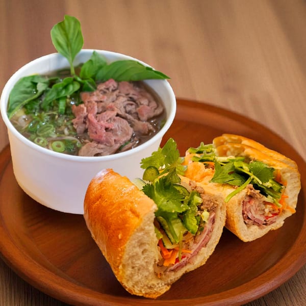 Vietnamese pho with beef and herbs in a white bowl, next to a banh mi sandwich.
