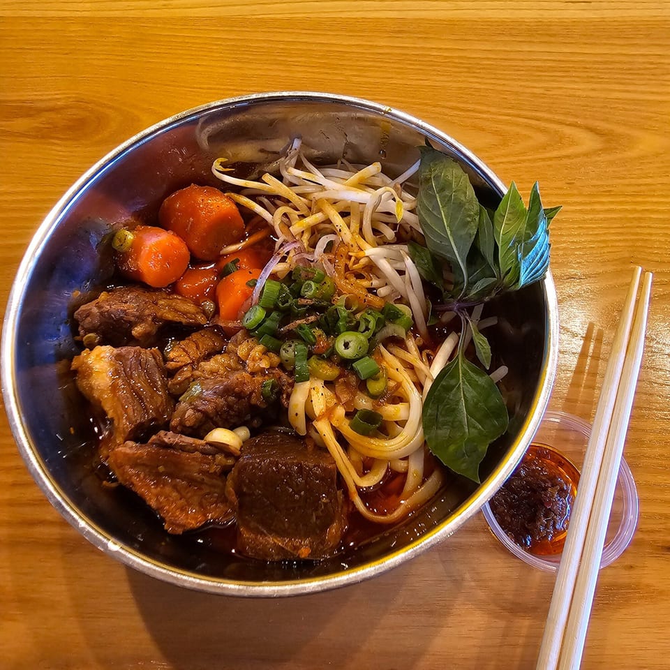 A bowl of Vietnamese beef stew with noodles, carrots, bean sprouts, and basil, with chopsticks and chili oil.