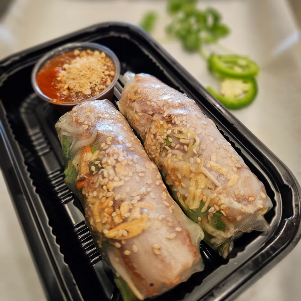 Two Vietnamese spring rolls with sesame seeds and a side of peanut dipping sauce in a black container.