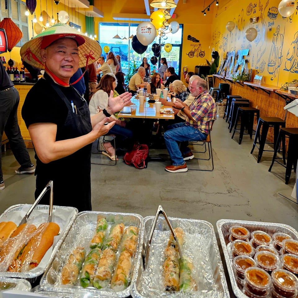 A man in a conical hat stands by trays of Vietnamese food, with diners in the background.