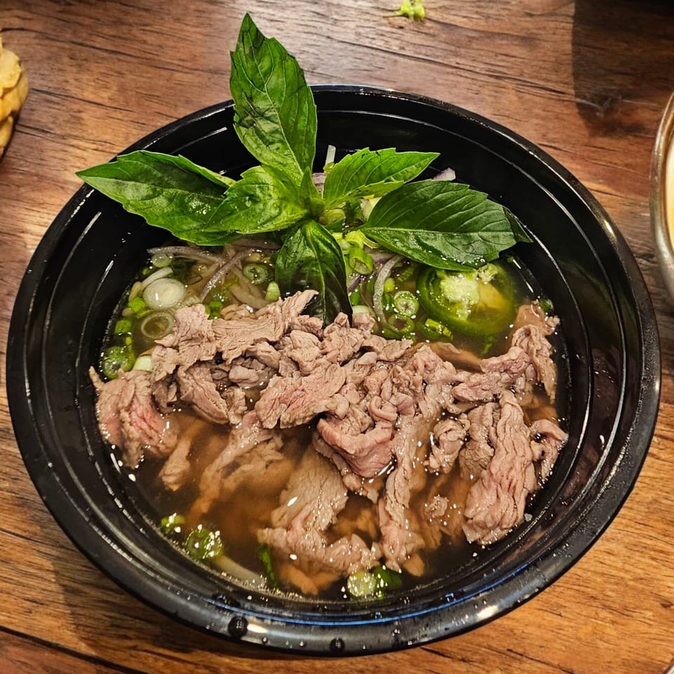 Vietnamese pho with beef, green onions, jalapeños, and basil in a black bowl on a wooden table.