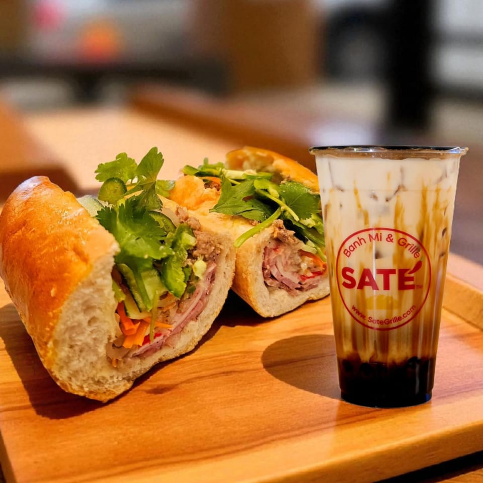 Two SATE Banh Mi sandwiches with fresh herbs and a SATE branded iced coffee on a wooden tray.