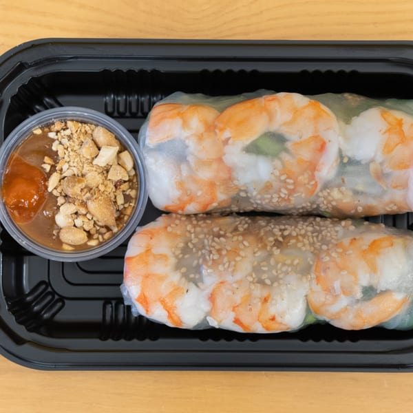 Two Vietnamese shrimp spring rolls with sesame seeds and peanut dipping sauce in a black container.
